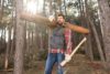 Un uomo nella foresta che trasporta un tronco sulla spalla destra e tiene un'ascia nella mano sinistra.