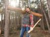 Un uomo nella foresta che trasporta un tronco sulla spalla destra e tiene un'ascia nella mano sinistra.